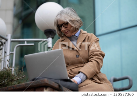 Senior woman working on laptop outdoors while sitting on steps near modern building during a sunny 120557360