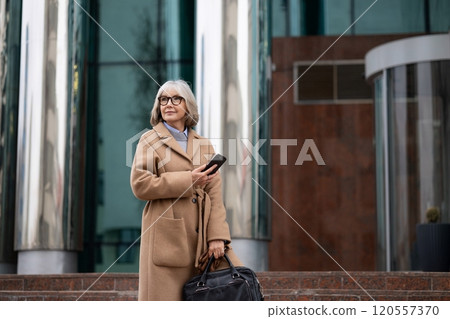 Older woman in a stylish coat stands outside a modern building holding a phone and bag on a sunny Older woman in a stylish coat stands outside a modern building holding a phone and bag on a sunny 120557370