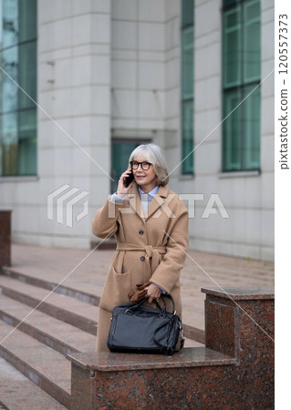 Senior woman wearing a stylish coat talks on her phone while standing on stone steps outside a Senior woman wearing a stylish coat talks on her phone while standing on stone steps outside a 120557373