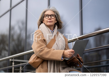 A senior woman sits outside a modern building in winter attire, working on her laptop while enjoying 120557390