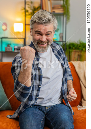 Happy Caucasian mature man watching TV shouting, celebrating success, winning, goal achievement Happy Caucasian mature man watching TV shouting, celebrating success, winning, goal achievement 120559590