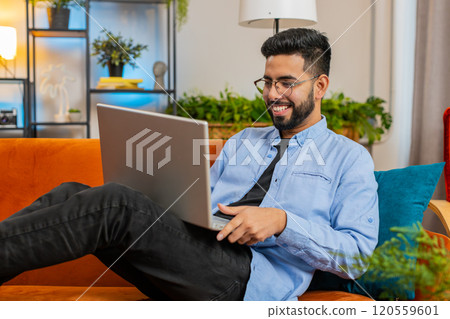 Indian man typing working on laptop using apps browsing internet lying study education on home sofa 120559601