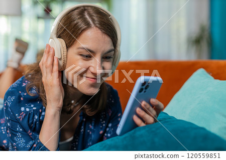 Young woman in wireless headphones relaxing lying on couch at home using smartphone listening music Young woman in wireless headphones relaxing lying on couch at home using smartphone listening music 120559851