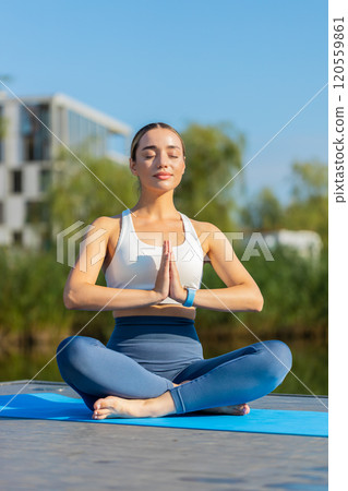 Caucasian young woman sitting on mat in lotus position, relaxing near pond in park on summer day 120559861