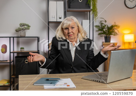 Confused senior businesswoman shrugging shoulders having doubts, no answer at home office desk Confused senior businesswoman shrugging shoulders having doubts, no answer at home office desk 120559870