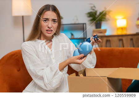 Dissatisfied annoyed young woman opening cardboard box receive broken wireless headphones at home 120559909