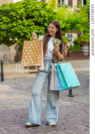Happy young shopaholic Caucasian woman holding euro cash while carrying shopping bags on city street Happy young shopaholic Caucasian woman holding euro cash while carrying shopping bags on city street 120559934