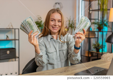 Excited businesswoman freelancer holding fan of dollar cash and dancing sitting at home office table 120559965
