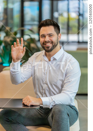 Portrait of smiling young businessman with laptop wave hand at camera sitting in hotel lobby 120560000