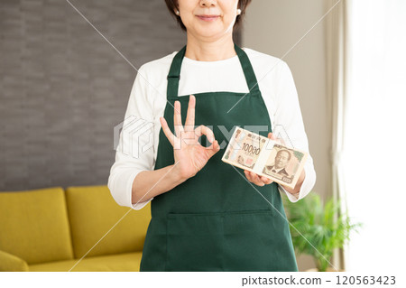 A middle-aged to senior woman housewife wearing a green apron making a circle sign while holding a stack of 1 million yen bills A middle-aged to senior woman housewife wearing a green apron making a circle sign while holding a stack of 1 million yen bills 120563423