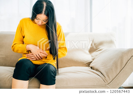 Highlighting severe stomachache, Woman seated on sofa suffers from intense abdominal pain. Emphasizing discomfort symptoms and the need for abdominal care. Health care abdomen bloating concept 120567443