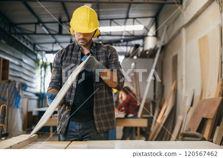 Construction worker in a yellow hardhat and safety glasses inspecting a board of wood. Ideal for carpentry, woodworking, and quality control, highlighting craftsmanship in the workshop. Labor day Construction worker in a yellow hardhat and safety glasses inspecting a board of wood. Ideal for carpentry, woodworking, and quality control, highlighting craftsmanship in the workshop. Labor day 120567462