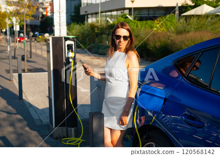 Woman in sunglasses and a fashionable white dress is using her smartphone while charging an electric car in the street. Woman in sunglasses and a fashionable white dress is using her smartphone while charging an electric car in the street. 120568142
