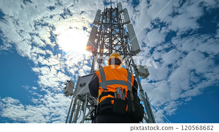 field technician inspecting cell tower under bright sky. technician wears safety vest and helmet, showcasing professional approach to telecommunications 120568682