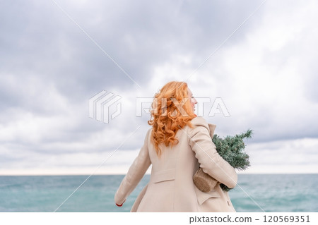Redhead woman Christmas tree sea. Christmas portrait of a happy redhead woman walking along the beach and holding a Christmas tree in her hands. Dressed in a light coat, white suit and red mittens. 120569351