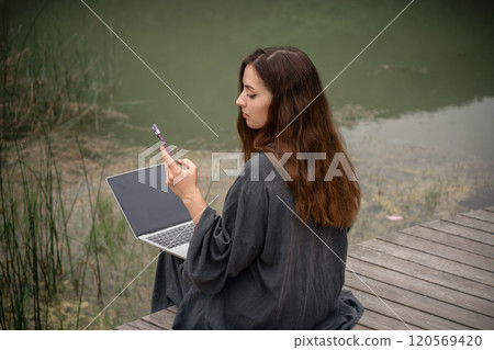 A woman is sitting on the ground with a laptop and a cell phone in her hands. She is using the laptop to browse the internet and the cell phone to communicate with someone. 120569420