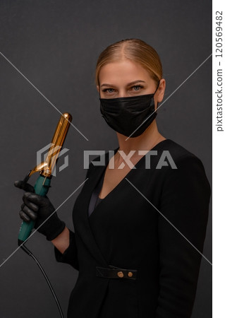 A woman in a black dress holding a hair curler. She is wearing a mask. Concept of caution and responsibility in the face of a pandemic. 120569482