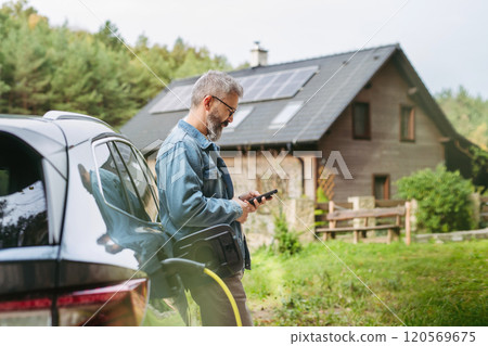 Man charging electric car in front of his house, plugging the charger into the charging port. House with solar panel system on roof behind him. Man charging electric car in front of his house, plugging the charger into the charging port. House with solar panel system on roof behind him. 120569675