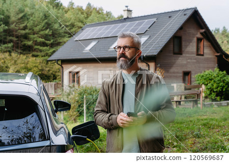 Man charging electric car in front of his house, plugging the charger into the charging port. House with solar panel system on roof behind him. Man charging electric car in front of his house, plugging the charger into the charging port. House with solar panel system on roof behind him. 120569687