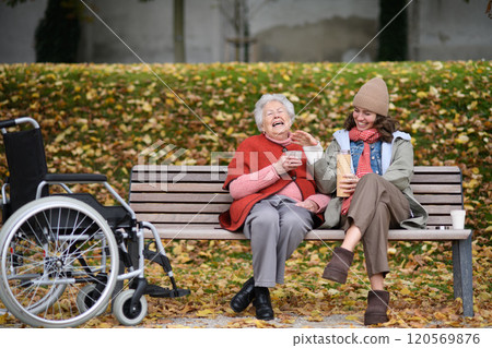 Portrait of a granddaughter and grandmother sitting on bench in autumn park, drinking coffee and laughing together. 120569876