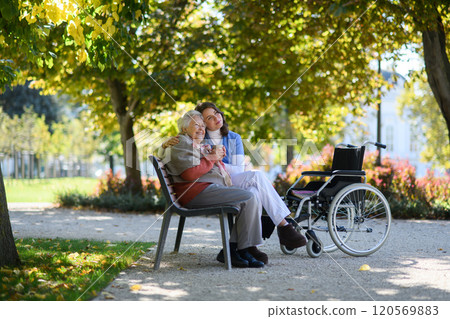 Elderly woman enjoying cup of coffee with her nurse in the park during warm autumn day. Young caregiver spending time with senior woman. 120569883