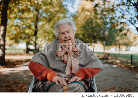 Portrait of elderly woman sitting in wheelchair, spending warm autumn day in city park Portrait of elderly woman sitting in wheelchair, spending warm autumn day in city park 120569895