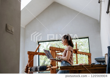 Young businesswoman working on laptop on workbench, standing by wooden loom. Upcycled textile, small weaving business and circular economy. 120569904