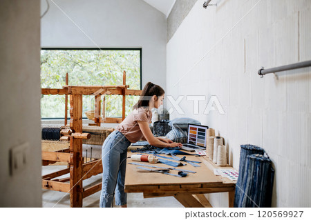Young businesswoman working on laptop on workbench, standing by wooden loom. Upcycled textile, small weaving business and circular economy. 120569927