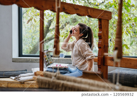 Young businesswoman making a call with customer, sitting by wooden loom. Upcycled textile, small weaving business and circular economy. 120569932