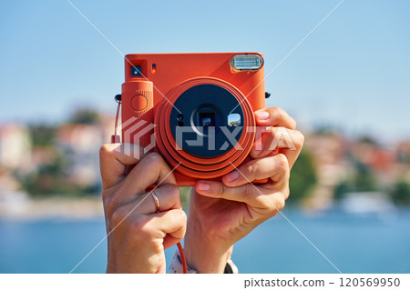 Close-up of tourist hand holding an orange instant camera in retro style with coastal town and blue sea in background. Summer travel concept. Equipment for capturing moments Close-up of tourist hand holding an orange instant camera in retro style with coastal town and blue sea in background. Summer travel concept. Equipment for capturing moments 120569950