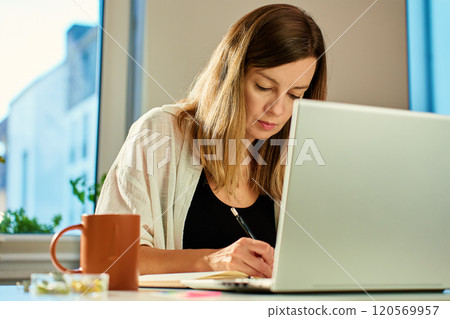 Freelance woman sitting at table using laptop and writing in notebook. Remote work at home office 120569957