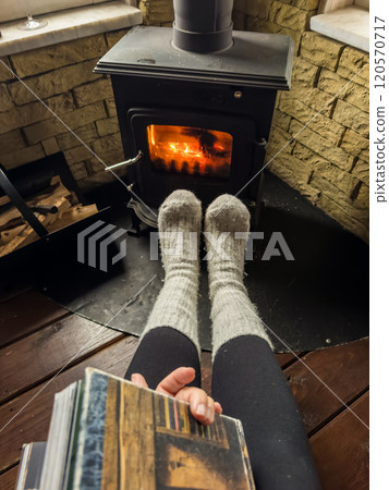 Woman relaxes by warm fire with book and warming up her feet in woollen socks. . High quality photo 120570717