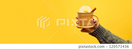 Banner. Hand dressed knitted sweater holding mug of hot, warm drink against warm yellow studio background with negative space to insert your. Banner. Hand dressed knitted sweater holding mug of hot, warm drink against warm yellow studio background with negative space to insert your. 120571436