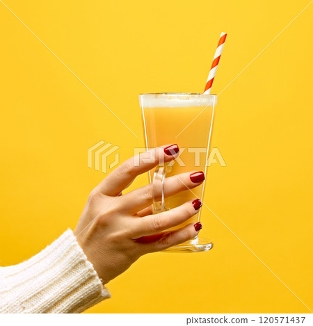 Hand in warm, knitted sweater holding glass of hot Toddy with red-white straw against warm yellow studio background. Hand in warm, knitted sweater holding glass of hot Toddy with red-white straw against warm yellow studio background. 120571437