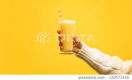 Hand in warm, knitted sweater holding glass of hot mead with red-white straw against warm yellow studio background. Hand in warm, knitted sweater holding glass of hot mead with red-white straw against warm yellow studio background. 120571438