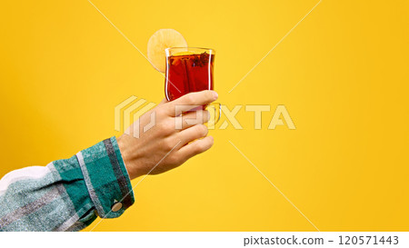 Woman's hand holds glass of hot mulled wine, garnished with apple slice and cinnamon stick, against warm yellow studio background. Woman's hand holds glass of hot mulled wine, garnished with apple slice and cinnamon stick, against warm yellow studio background. 120571443