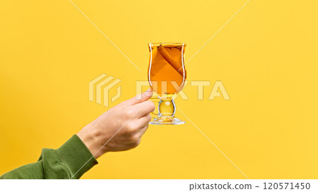 Woman's hand holds glass of punch garnished with citrus slice and cinnamon stick, against warm yellow studio background. Woman's hand holds glass of punch garnished with citrus slice and cinnamon stick, against warm yellow studio background. 120571450