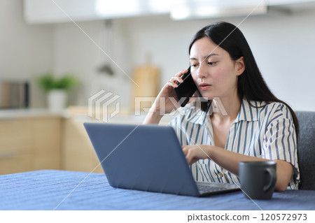 Asian woman talking on phone checking laptop solving problem Asian woman talking on phone checking laptop solving problem 120572973
