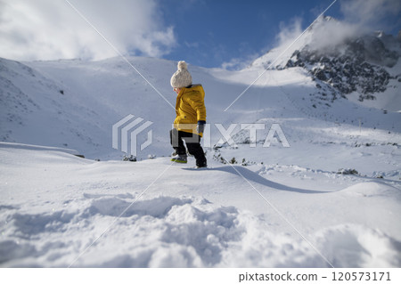 Portrait of boy in yellow jacket walking across snowy nature, enjoying winter holiday in the mountains with family. 120573171