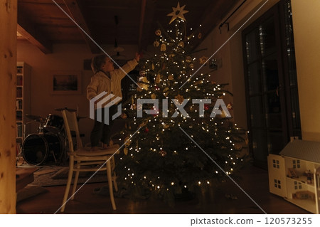 Portrait of cute boy standing on chair and helping with decorating the Christmas tree with ornaments. Portrait of cute boy standing on chair and helping with decorating the Christmas tree with ornaments. 120573255