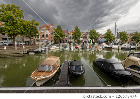 Serene harbor with moored boats under a dramatic cloudy sky. Quaint historic buildings and trees line the waterfront, reflecting in the calm green water, creating peaceful scene. Dordrecht landscape. 120575474