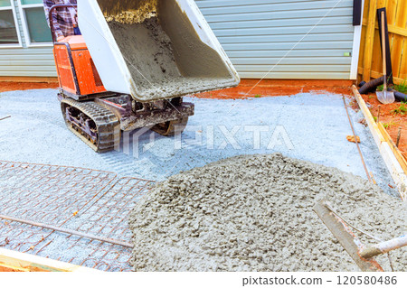 Heavy machinery dispenses fresh concrete onto prepared area for new patio workers guide process in suburban backyard using compact tracked dumper 120580486