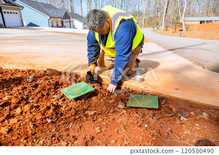 Worker wearing safety vest kneels on ground while inspecting maintaining drainage system outside several homes. Worker wearing safety vest kneels on ground while inspecting maintaining drainage system outside several homes. 120580490