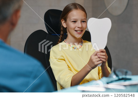 Smiling Girl Holding Mirror While Sitting in Dental Chair Smiling Girl Holding Mirror While Sitting in Dental Chair 120582548