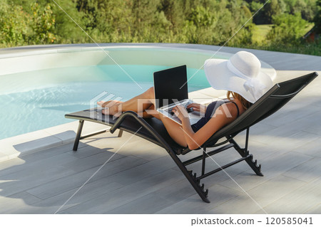 Woman working on laptop by poolside outdoors 120585041