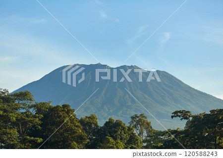 Panorama of Mount Agung and rice fields on the island of Bali. View of the mountain against a background of palm trees and a cornfield. Panorama of Agung volcano covered with clouds on a sunny day. Panorama of Mount Agung and rice fields on the island of Bali. View of the mountain against a background of palm trees and a cornfield. Panorama of Agung volcano covered with clouds on a sunny day. 120588243