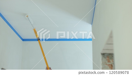 Man makes repairs inside the house, painting the ceiling with a long stick with a roller. Home repairs and finishing works. 120589078