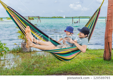 Father with his toddler son lying in a hammock against the backdrop of turquoise waters of Bacalar Lake, Mexico. Peaceful tropical travel destination concept 120589765