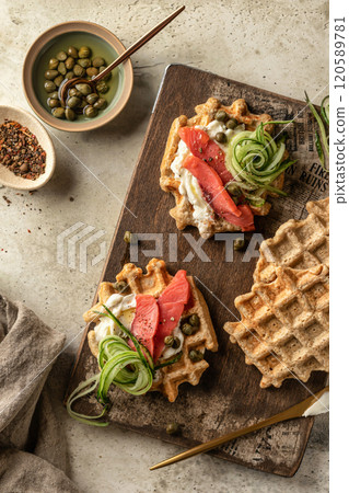 Savory waffles. Breakfast waffles with cream cheese, cucumber, salmon on wooden board for breakfast on rustic textured background. Top view Savory waffles. Breakfast waffles with cream cheese, cucumber, salmon on wooden board for breakfast on rustic textured background. Top view 120589781