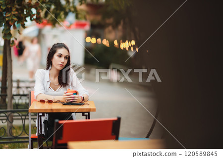 Young woman sitting at outdoor cafe table with colorful mug in hand Young woman sitting at outdoor cafe table with colorful mug in hand 120589845
